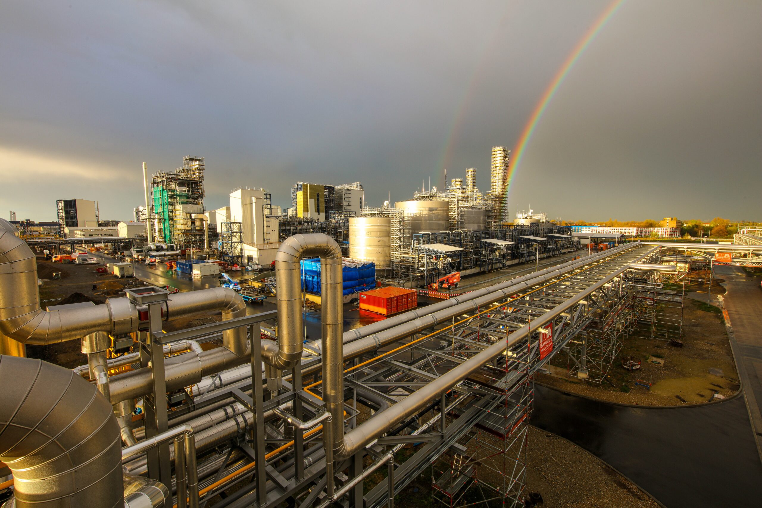 Baustellenkamera-Vorteile: Überwachung einer großen Industriebaustelle mit modernen Kamerasystemen. Zu sehen sind Rohre, Gerüste und Gebäude im Bau, während zwei Regenbögen den Himmel über der Baustelle schmücken. Die Kamera ermöglicht Echtzeitüberwachung, lückenlose Dokumentation und effizientes Projektmanagement.