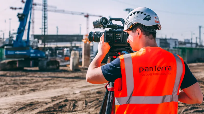 panTerra Kameramann mit Helm und Warnweste filmt mit Videokamera auf Stativ auf einer Baustelle